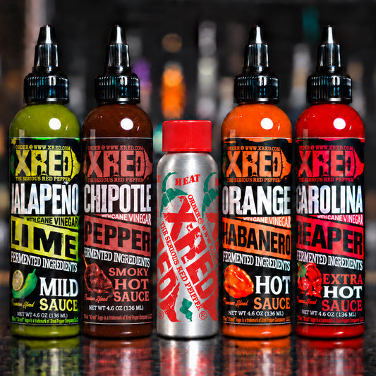 Five bottles of Xred Hot Sauce arranged in a row on a dark countertop, with blurred bar shelves in the background. The bottles include Jalapeño Lime, Chipotle Pepper, Smokez Hot Sauce, Habanero Reaper, and Carolina Reaper.