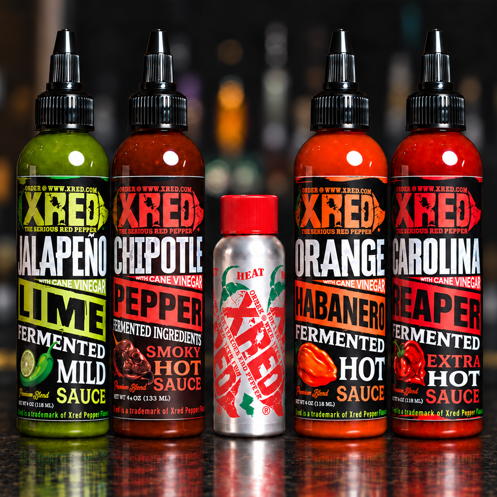Five bottles of Xred Hot Sauce products lined up on a dark bar counter with a blurred bar background. The lineup includes a green Jalapeño Lime Fermented Mild Sauce bottle, a red Chipotle Pepper Fermented Ingredient bottle, a silver Xred Hot Sauce bottle with a red cap, and two red Carolina Reaper Fermented Extra Hot Sauce bottles.